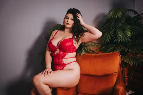 Curvy woman in red lace lingerie sitting on an orange velvet armchair in a moody indoor setting.