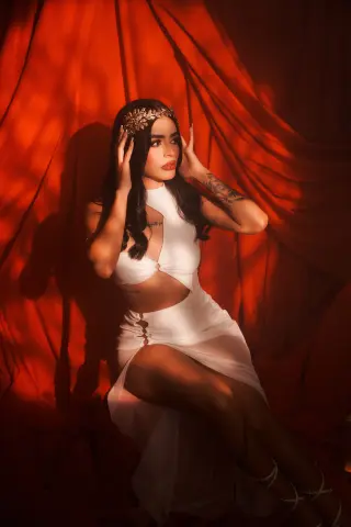 Elegant woman in a white goddess dress and gold leaf crown posing against red velvet curtains.