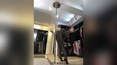 NicoleBella arches her back while standing next to a silver pole, wearing a black sheer bodysuit and black high heels.