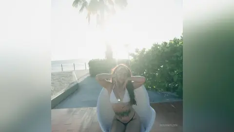 RossiNoleen poses in a white bikini top and black bottoms while sitting outdoors near palm trees and a beach view.