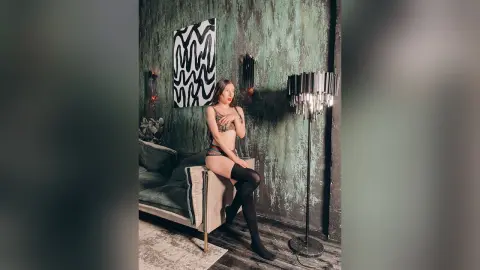 Young woman in black lingerie and stockings posing on a velvet sofa in a moody, dark green aesthetic room.