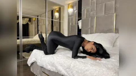 Woman in a black bodysuit lounging on a white faux fur bed in a modern luxury bedroom.