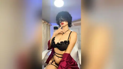 Glamorous woman in black lace lingerie and a masquerade mask posing in a bright indoor setting.