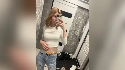 Young woman in a white ribbed long-sleeve top and blue jeans taking a mirror selfie in a modern room.