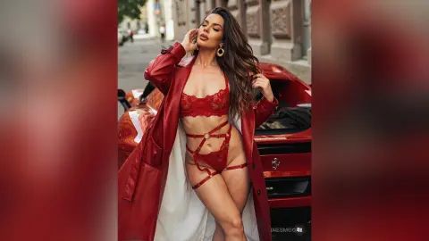 InesMoore poses in red lace lingerie and a long red coat, leaning against a red car on a city street with her long brown hair down.