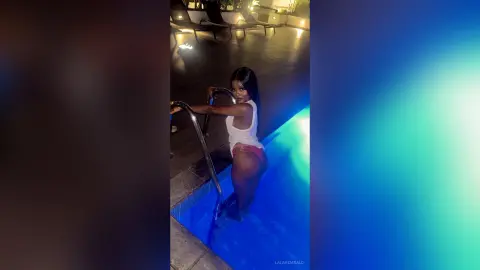 Young woman in a white tank top and bikini relaxing in a glowing blue swimming pool at night.