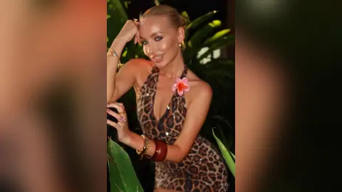 Smiling blonde woman in a leopard print halter dress posing amidst tropical greenery for a summer vacation vibe.