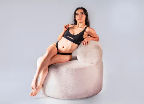 Young woman in black bikini swimwear sitting on a white boucle chair against a plain studio background.