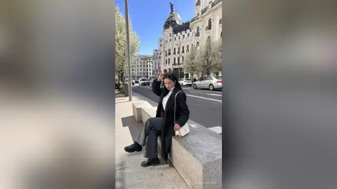 NashiroSakurai sits on a stone ledge wearing a black coat, white shirt, and grey trousers against a city street background.