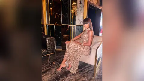 LanaRhode sits on a white chair wearing a sparkly, tight halter dress and high heels in a luxury room.