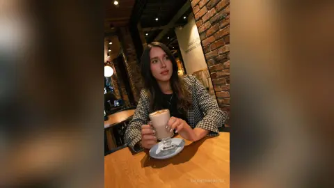 ValentinaGarcez sits at a wooden table in a cafe, wearing a black top and a black and white plaid blazer while holding a latte in a glass mug.