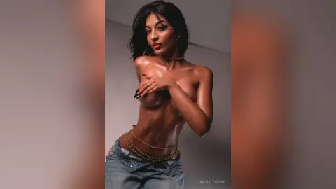 Glamorous woman with glowing skin wearing denim jeans and gold body chains in a moody studio setting.