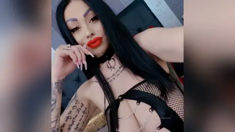DeeaAnderson poses with long black hair and red lipstick, wearing a black mesh choker and black fishnet lingerie.