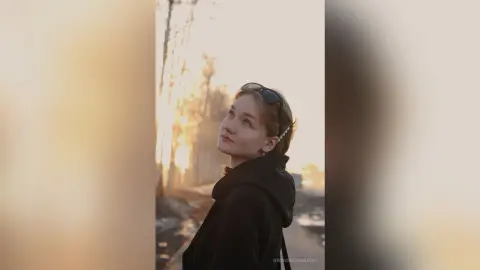 GwendolynBucey looks over her shoulder wearing a black hoodie and sunglasses with her hair pulled back against a sunset background.