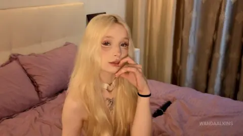 Blonde woman in a white top sitting on a purple bed in a cozy, soft-lit bedroom setting.