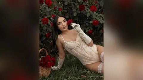 Beautiful woman in cream lace lingerie and white gloves holding a red rose in a romantic rose garden.