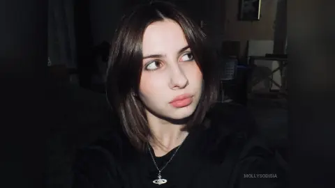 Moody portrait of a young woman with brown hair wearing a black shirt and silver necklace in dim lighting.