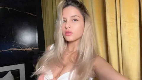 Blonde woman in a white satin top posing against yellow curtains in a moody, elegant indoor setting.