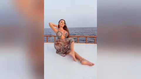 Glamorous woman in a patterned cutout maxi dress relaxing on a boat deck overlooking the ocean at sunset.