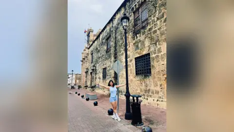 Cheerful woman in a light blue tank top and denim shorts standing by a vintage lamp post on a cobblestone street.