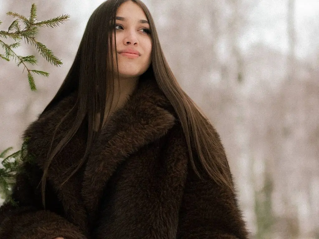 Young woman in a cozy brown faux fur coat posing in a dreamy, misty winter forest setting.