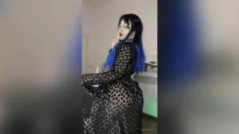 Gothic woman with blue hair in a black sheer leopard print dress posing in a moody indoor setting.