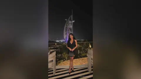 Elegant woman in a black cocktail dress posing on a luxury balcony with the Burj Al Arab at night.