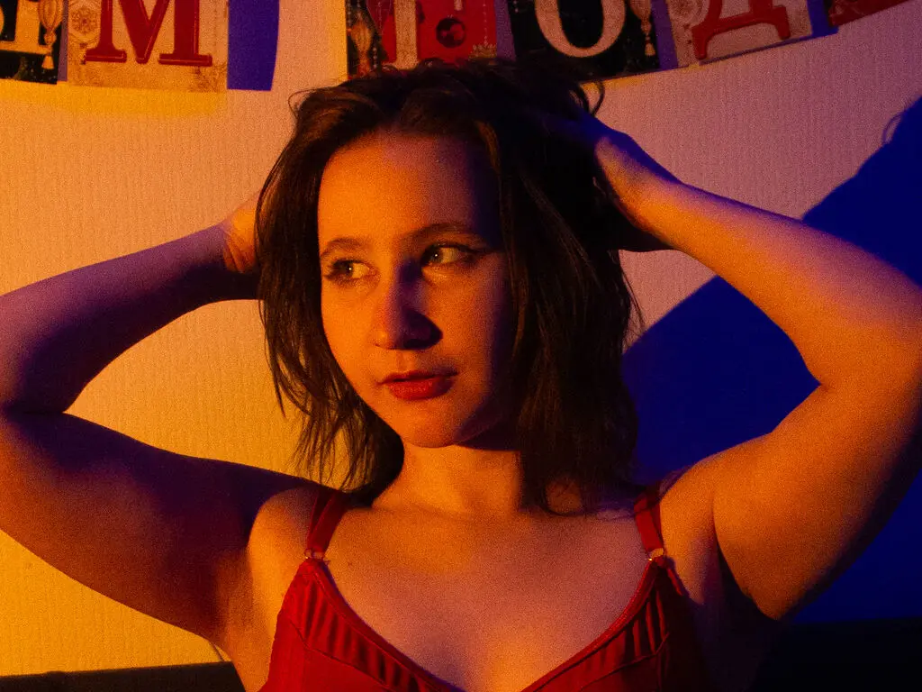 ChristyWeb poses with her hands in her dark hair, wearing a red camisole against a warm, lit wall.