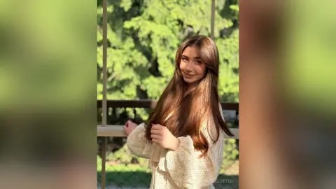 Smiling young woman with long brown hair in a white lace blouse posing in a sunny garden setting.