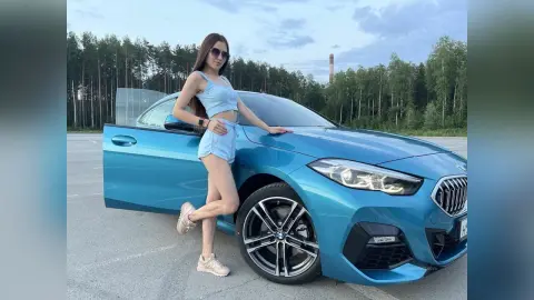 MilanaVolkova poses next to a blue car, wearing a light blue crop top and matching shorts with her long brown hair down.