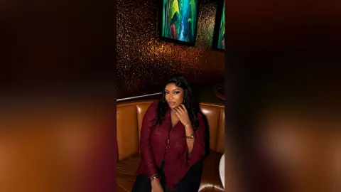 RubyJavis poses in a dark setting wearing a deep red lace top, sitting in a brown leather booth with her hand near her face.