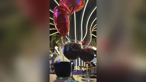 Two glasses of red wine on a table with festive pink balloons in a dimly lit, moody restaurant setting.