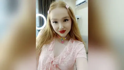 Smiling blonde woman in a pink lace top and colorful beaded necklace posing in a bright room.