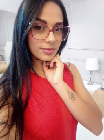 Young woman with long dark hair wearing pink glasses and a red lace top in a bright bedroom setting.