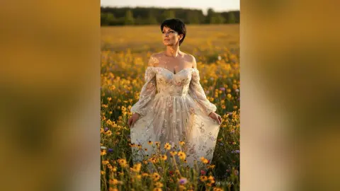 Elegant woman in a floral lace gown standing in a golden wildflower meadow at sunset.