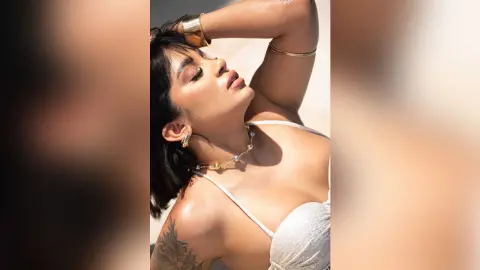 Glamorous woman in a white bikini and gold jewelry posing under bright sunlight at a sunny beach.