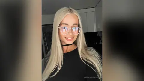 Smiling blonde woman with glasses and black choker in a modern indoor setting.