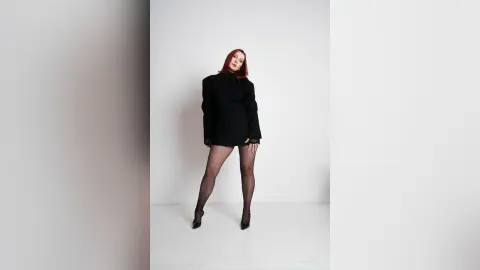 AryaSin poses in a black long-sleeved mini dress and sheer black tights against a plain white background.