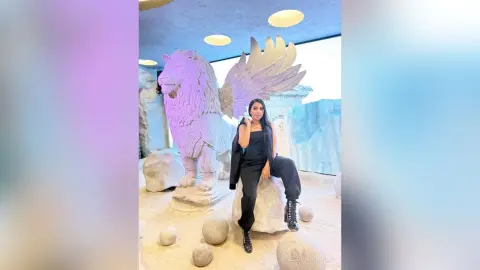 Woman in black jumpsuit sitting by a large white winged lion statue in a dreamy, ethereal indoor setting.