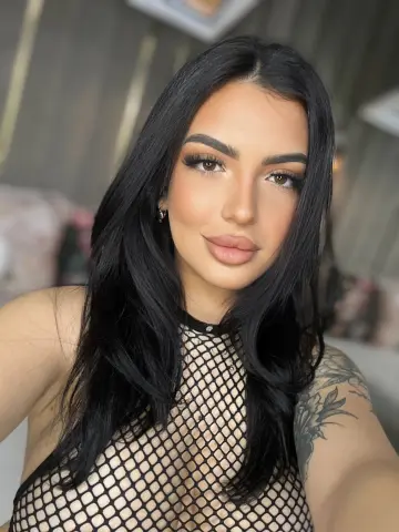 NoraSin poses with long black hair wearing a black fishnet top that shows her cleavage and shoulder tattoo.