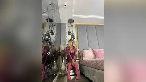 NicoleBella poses in a pink lace bra and matching pink trousers while sitting on a grey velvet bed.