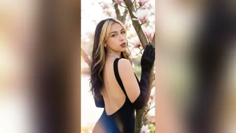 Elegant woman in a backless black dress and long gloves posing amidst blooming pink flowers in a dreamy garden.