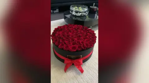 A large, round black box filled with a dense arrangement of deep red roses, tied with a red ribbon bow.