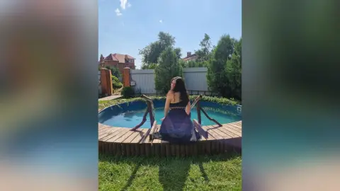 LateshaKaspar sits on a wooden deck by a swimming pool, wearing a dark tank top and looking back over her shoulder with her long dark hair down.