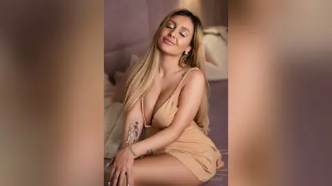 Anita poses on a bed wearing a tan silk slip dress, showing off her long blonde hair and arm tattoos.