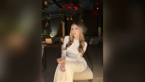 Elegant woman in a white long-sleeve outfit sitting in a moody, dimly lit upscale lounge setting.