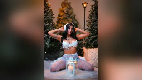 Aleja poses nakedly in white lace lingerie, sitting on a white furry surface surrounded by lit Christmas trees.