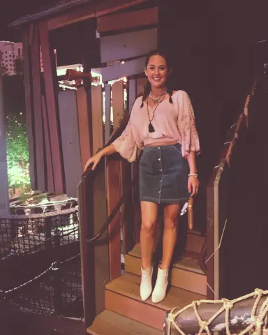 Smiling woman in a pink blouse and denim skirt standing on a wooden balcony at night.