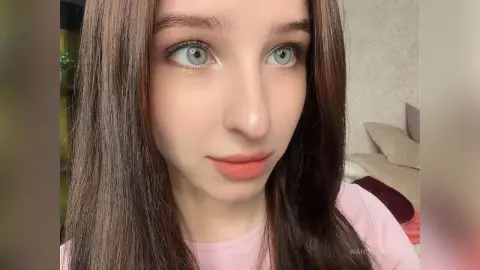 WanitaCruson has long brown hair and light eyes, wearing a pink shirt while looking off-camera in a room with a bed behind her.