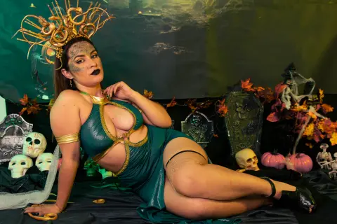 Woman in dark green goddess costume and gold crown posing in a spooky Halloween setting with skulls and pumpkins.